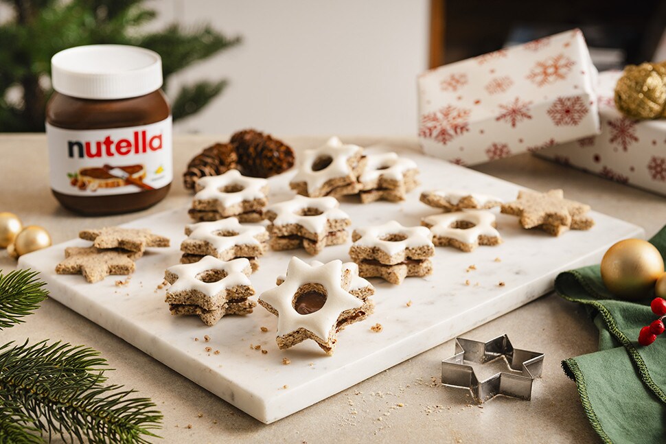 Mehrere frisch gebackene und glasierte Zimtsterne liegen auf einer weißen Marmorplatte. Neben der Marmorplatte steht ein Glas nutella®.