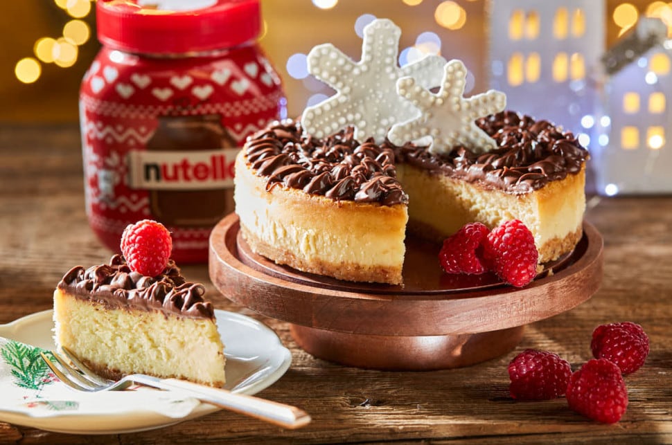 Nutella® Mascarpone Cheesecake est présenté sur un présentoir à gâteaux en bois. Une part de gâteau a été découpée et est posée sur une assiette à gauche du gâteau.
