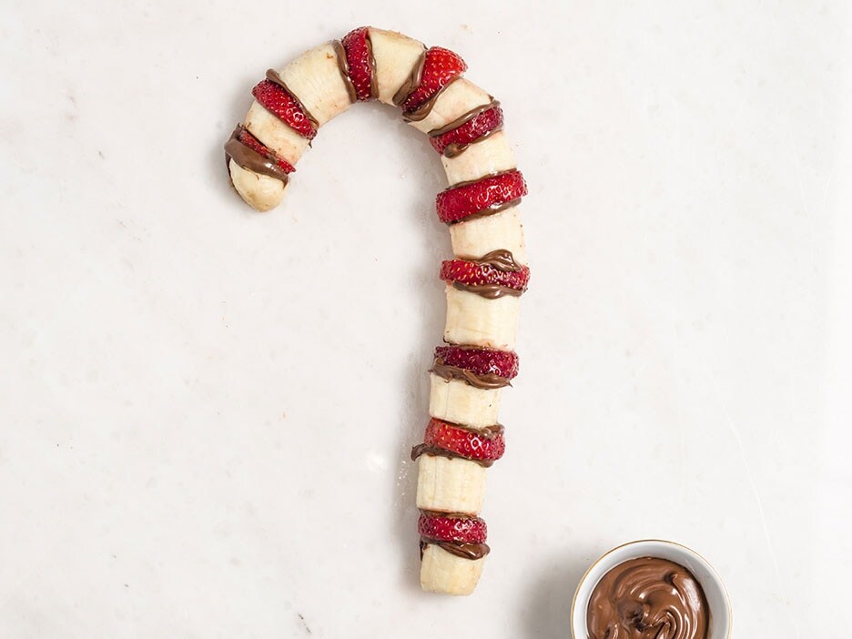 Candy-cane-shaped fruit skewers dipped in Nutella, arranged on wooden board.
