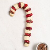 Candy-cane-shaped fruit skewers dipped in Nutella, arranged on wooden board.