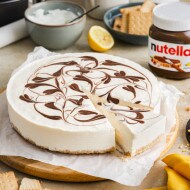 A no bake cheesecake with swirls of Nutella® on top is displayed on a circular wooden chopping board surrounded by a jar of Nutella® and other no bake cheesecake ingredients.
