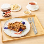 Slices of banana bread topped with Nutella® are arranged on a plate resting on a breakfast tray. In the background, a jar of Nutella® and a cup of tea.