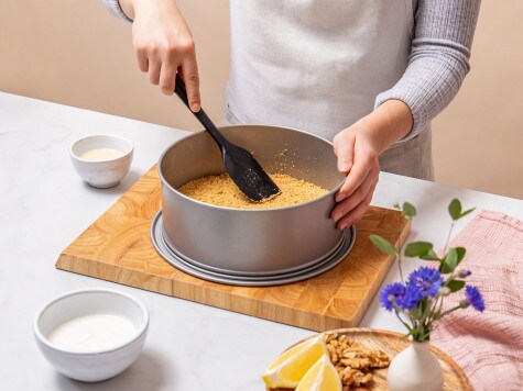 A baker is spreading the crumb mixture into a springform pan and pressing it into a smooth base.