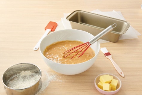 The ingredients for the banana bread are in a large bowl with a whisk. In the background, a rectangular cake pan.