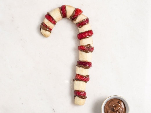 Candy-cane-shaped fruit skewers dipped in Nutella, arranged on wooden board.