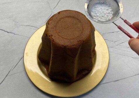 Il Pandoro è adagiato su un piatto d'oro. Un piccolo setaccio contenente zucchero a velo è sospeso sopra la torta.