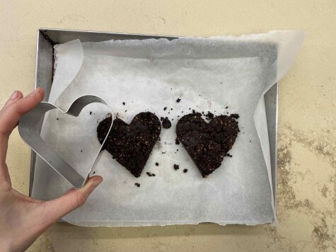 Vista dall'alto di una teglia foderata con carta da forno con due crumble a forma di cuore posizionati al centro. Una mano sta sollevando lo stampo a forma di cuore dal crumble.