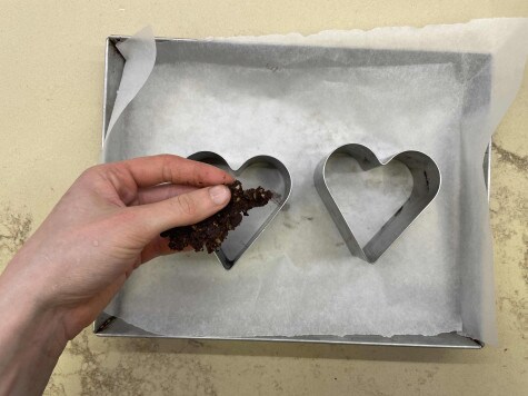 Vista dall'alto di una teglia foderata con carta da forno con due stampi a forma di cuore posizionati al centro. Una mano sta riempiendo gli stampi con lil crumble.