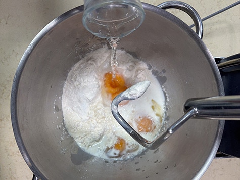 Vista dall'alto di una ciotola contenente farina e uova. L'acqua viene versata da un barattolo di vetro.
