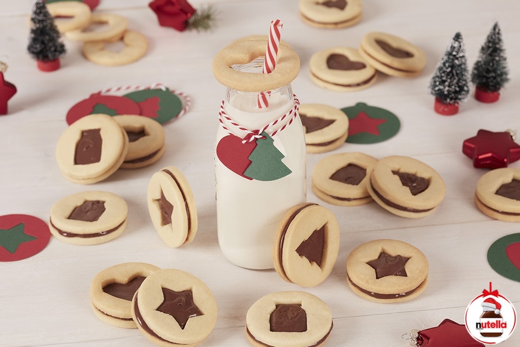 A milk bottle with a red-striped straw is surrounded by star and tree-shaped chocolate-filled cookies on a wooden table. Red and green paper tags hang from the bottle.