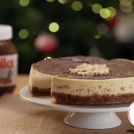 A cheesecake with a chocolate topping sits on a white pedestal. In the background, a jar labeled "Nutella" is visible, with blurred holiday lights creating a festive atmosphere.