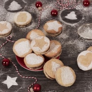 Cookies with powdered sugar are decorated with holiday shapes and arranged on a table. Red baubles and twine are scattered around, and similar patterns appear dusted on the dark surface.