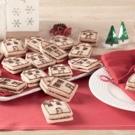 Cookies shaped like houses are piled on a plate and table, decorated with brown icing. A festive setting includes a red cloth, white plates, and small green tree ornaments.