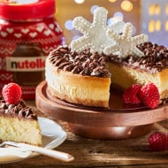 Nutella® Mascarpone Cheesecake is presented on a wooden cake stand. A slice of cake has been cut and is placed on a plate to the left of the cake.