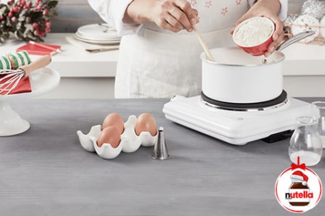 A baker adds flour from a small ceramic bowl into a pot while stirring the contents in the pot with a wooden spoon.