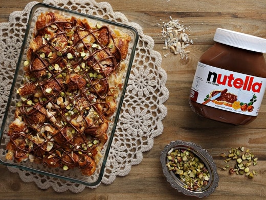 A baked dessert topped with pistachios and Nutella drizzle sits in a glass dish on a lace mat. Nearby, a jar of Nutella, almond slivers, and a dish of pistachios rest on a wooden table.