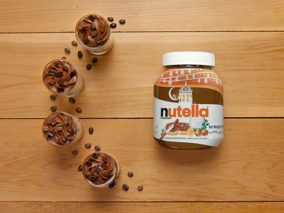 جرة نوتيلا موضوعة على سطح خشبي بجانب أربعة أكواب حلوى صغيرة مملوءة بموس الشوكولاتة، ومحاطة بحبوب القهوة المتناثرة.