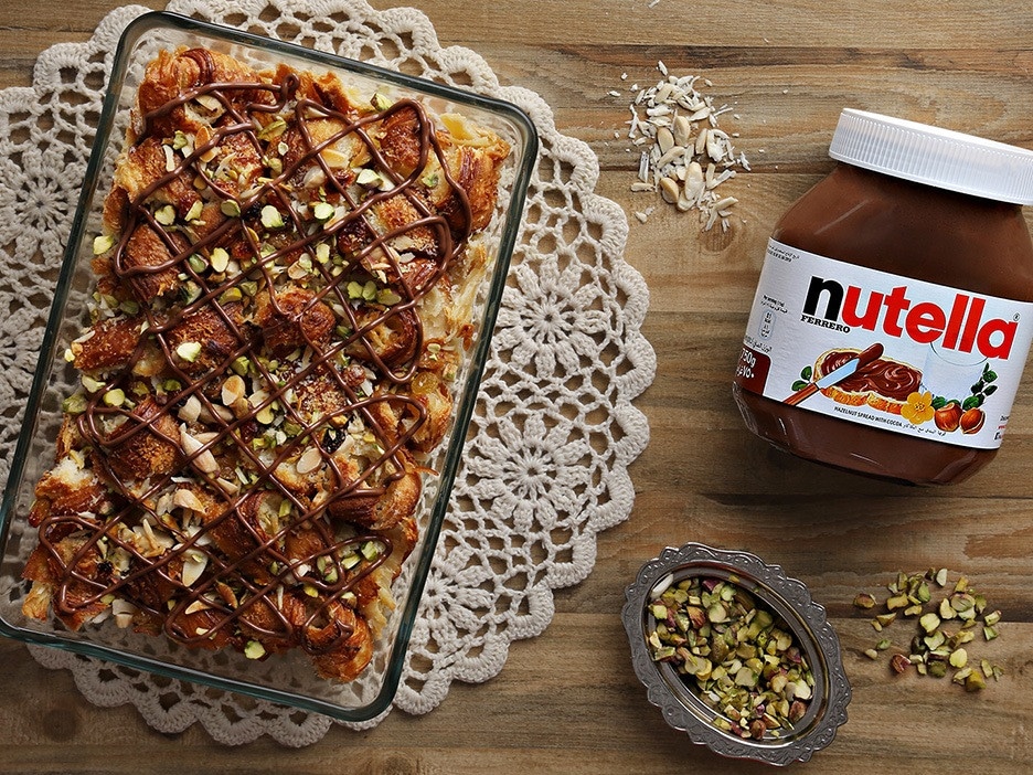 A baked dessert topped with pistachios and Nutella drizzle sits in a glass dish on a lace mat. Nearby, a jar of Nutella, almond slivers, and a dish of pistachios rest on a wooden table.