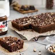 El brownie de Nutella® se coloca sobre una rejilla forrada con papel para hornear para que se enfríe. Del brownie se cortó un cuadrado y se colocó junto a un frasco de Nutella®.