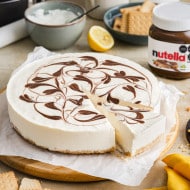Una tarta de queso sin hornear con rizos de Nutella® encima se muestra sobre una tabla de cortar circular de madera rodeada por un frasco de Nutella® y otros ingredientes de tarta de queso sin hornear.