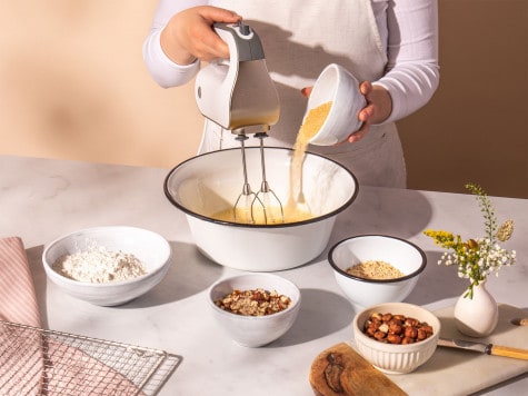 Un repostero sostiene un batidor en un tazón grande y vierte la masa en él. Pequeños tazones con ingredientes para el brownie rodean el tazón.