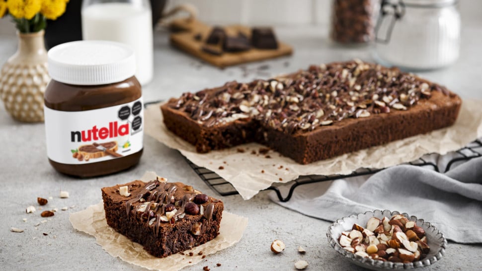 El brownie de Nutella® se coloca sobre una rejilla forrada con papel para hornear para que se enfríe. Del brownie se cortó un cuadrado y se colocó junto a un frasco de Nutella®.