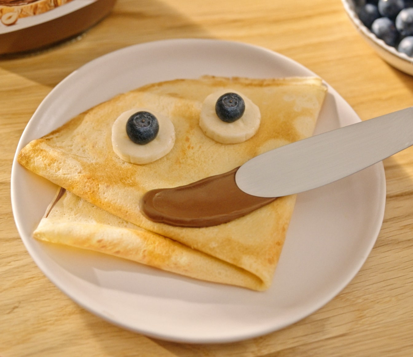 Um crepe dobrado num prato branco, decorado com Nutella, rodelas de banana e mirtilos para formar uma cara sorridente.