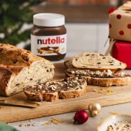 A festive loaf of Christmas Stollen sliced on a wooden board, topped with creamy Nutella® and chopped nuts. A jar of Nutella®, wrapped gifts, and Christmas decorations sit in the background: