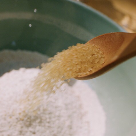 A wooden spoon pouring raw brown sugar into a mixing bowl filled with white flour, captured mid-motion.