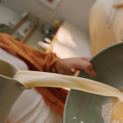 A person pouring soy milk from a jug into a mixing bowl partially filled with dry ingredients
