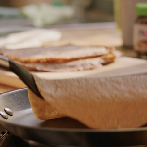A crepe is being cooked in a non-stick frying pan. A spatula is lifting the edge.