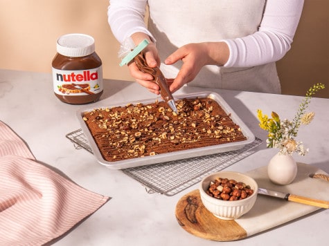 A baker decorates the baked brownie, which is then spread on a baking sheet with hazelnuts and Nutella®.