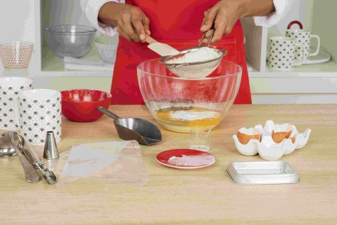 A baker is sifting flour into a glass mixing bowl containing eggs, milk, sugar and baking powder.