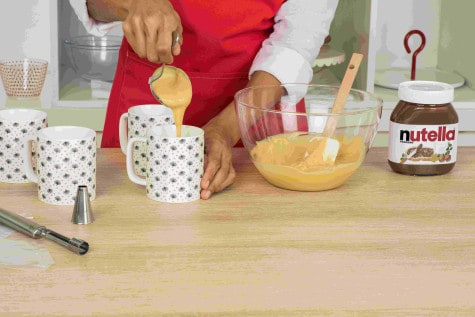 A baker is pouring mug cake mixture from a glass mixing bowl into 4 separate mugs.