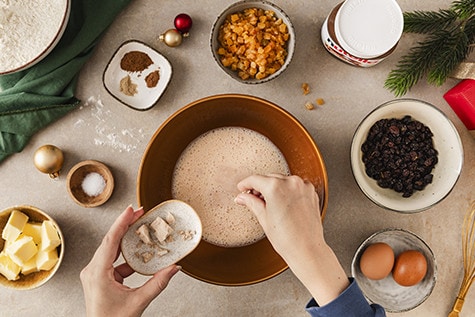 Hands preparing Christmas Stollen ingredients - adding fresh yeast to a bowl of warm milk mixture. Surrounding the bowl are butter cubes, flour, dried fruit, eggs, spices, salt, and a jar of Nutella®