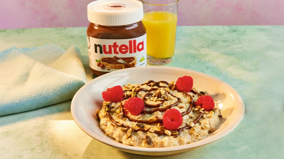 A bowl of porridge topped with toasted nuts, seeds, raspberries, and Nutella®, served with a Nutella® jar.