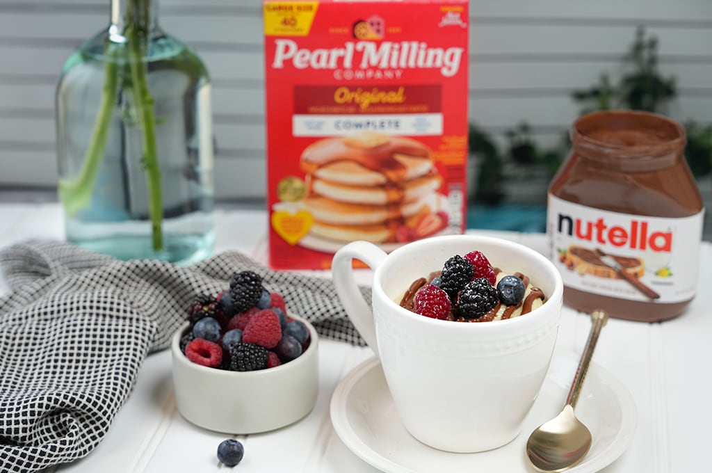 Mug Pancake with Nutella® and berries in a white mug and saucer, a black and white towel, some berries, Nutella® jar, and pearl milling company pancake mix nearby.