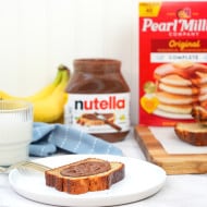 A slice of banana bread with Nutella® on a white plate, a fork, glass of milk, Nutella® jar, bananas and pearl milling company pancake mix are placed in the background.