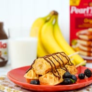 Banana On A Stick with Nutella® on a red plate surrounded by bunch of bananas, berries, a glass of milk, Nutella® jar, and Pearl Milling Company pancake mix.