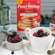 Mug Pancake with Nutella® and berries in a white mug and saucer, a black and white towel, some berries, Nutella® jar, and pearl milling company pancake mix nearby.