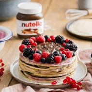 A Nutella® crepe cake, decorated with fresh berries is displayed on a plate. A jar of Nutella®, fresh berries and cream are shown in the background.