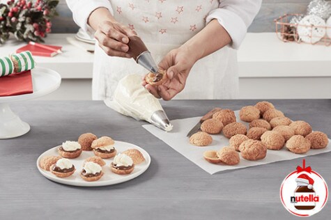 A baker is piping small dots of Nutella® into the center of cut in half cream puffs using a piping bag.