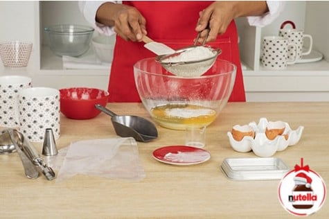 A baker is sifting flour into a glass mixing bowl containing eggs, milk, sugar and baking powder.