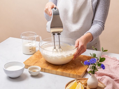 A baker is whipping cream in a glass mixing bowl using an electric whisk to make the no bake cheesecake filling.