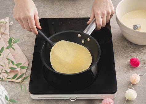 A crepe is being cooked in a small frying pan, with a silicone spatula underneath it to prevent it from sticking to the pan.