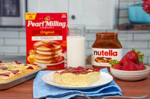 A slice of sheet pan pancake decorated with Nutella® on a white plate and blue napkin, surrounded by strawberries, glass of milk, Nutella® jar, and pearl milling company pancake mix.