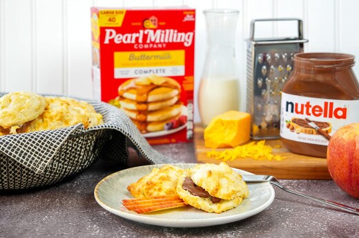 Apple Cheddar Biscuits with Nutella® and cut apples on a plate, Nutella® jar, cheese, more cheese biscuits and Pearl Milling Company buttermilk pancake mix are placed in the background.