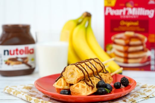 Banana On A Stick with Nutella® on a red plate surrounded by bunch of bananas, berries, a glass of milk, Nutella® jar, and Pearl Milling Company pancake mix.