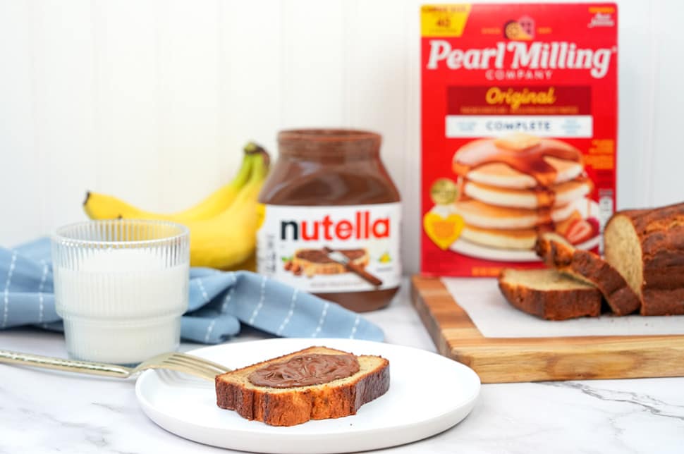 A slice of banana bread with Nutella® on a white plate, a fork, glass of milk, Nutella® jar, bananas and pearl milling company pancake mix are placed in the background.