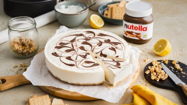 A no bake cheesecake with swirls of Nutella® on top is displayed on a circular wooden chopping board surrounded by a jar of Nutella® and other no bake cheesecake ingredients.